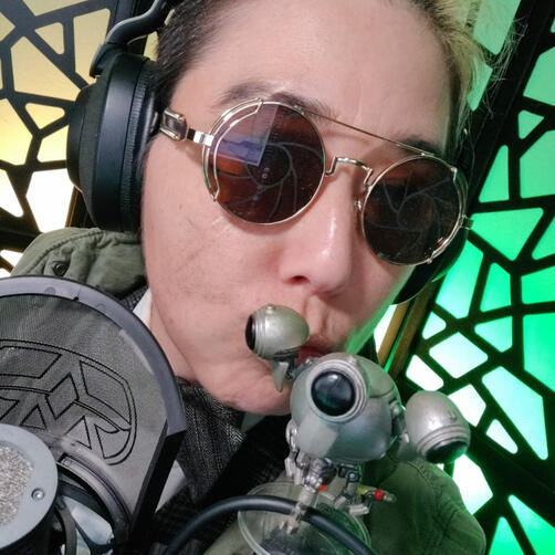 Cai leaning close in to the camera wearing large, round sunglasses with irising lenses drawn onto them. He is dressed as Mart from Angels & Atoms on Vault Companions.