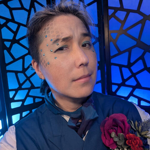 Matteo Cai dressed as Matteo from the Ghoul Society one-shot on RoleD5. He has a blue waistcoat with a blue ascot on. On top of it all he has a massive floral boutinere pinned to his waistcoat. He has scales painted onto his face with blue crystals.