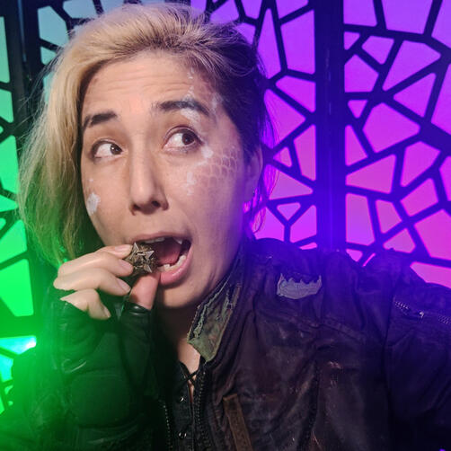 Cai as Rivers Ommerica from Roguelike Rumbles with Pick Your Poison Productions. They have dark scales painted on their face with white cracks paited over them. They are wearing distressed clothing with patches and rips in them and are miming about to eat