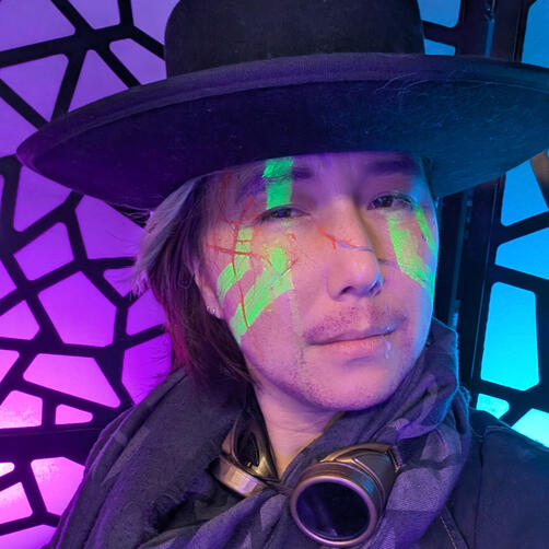 Xainan Cai as Xainan Esch from Transplanar RPG's The Chaos Protocol's Arc 3. Cai wears a black, flat-brimmed hat, a black leather motorcycle jacket and a black henley. Their shoulder-length hair falls only on one side of their face, lightly covering black cracks