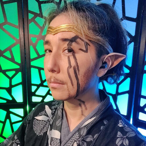 Hoshiro Cai dressed as Hoshiro from Sun in Chains on BadHouseRPG. He has his dark hair pulled back in a somewhat messy high ponytail and natural-looking makeup with a thin gotee. There is a black swirling face marking that goes from his left brow down past the nec