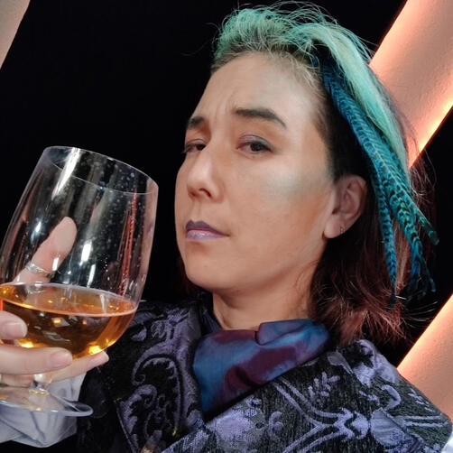 Rhode Theyolas Cai as Rhode from Outrunners on Happy Jacks RPG. He hais teal makeup including scales, hair color, and feathers. He raises a glass to the camera filled with amber liquid.