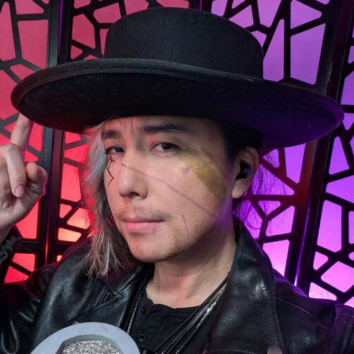 Xainan Cai as Xainan Esch from Transplanar RPG's The Chaos Protocol's Arc 3. Cai wears a black, flat-brimmed hat, a black leather motorcycle jacket and a black henley. Their shoulder-length hair falls only on one side of their face, lightly covering black cracks